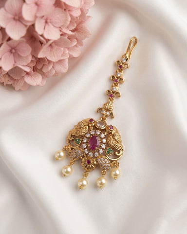 An ornate gold-toned Indian jhumka earring decorated with a central pinkish-red stone, small green and pink stones, clear pavé-like accents, and dangling pearl beads, displayed on white satin fabric with pink flowers in the corner.