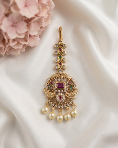 An ornate gold-toned traditional Indian pendant or earring decorated with a central ruby gemstone, surrounded by small clear stones, green and red accent stones, and five dangling pearl beads, displayed on white satin fabric with pink flowers in the corner.