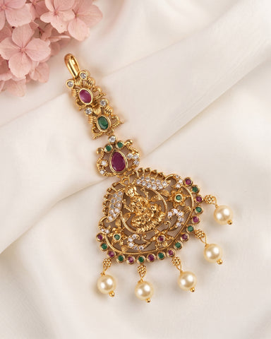 An ornate gold-toned traditional Indian jhumka earring decorated with red, green, and white stones, featuring dangling pearl-like beads, displayed on a draped white fabric background with pink flowers in the corner.