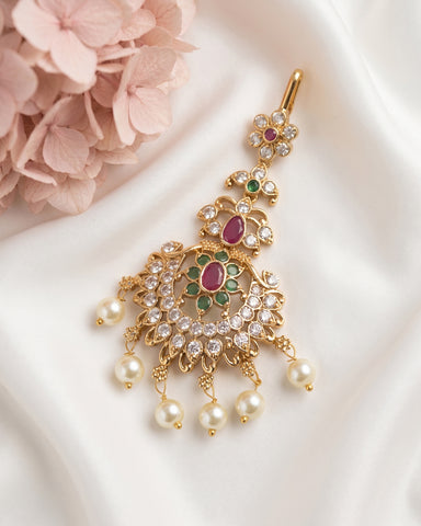 Gold-toned traditional Indian jhumka earrings with green and pink stones and white pearl drops, displayed on a white satin fabric next to pink flowers.