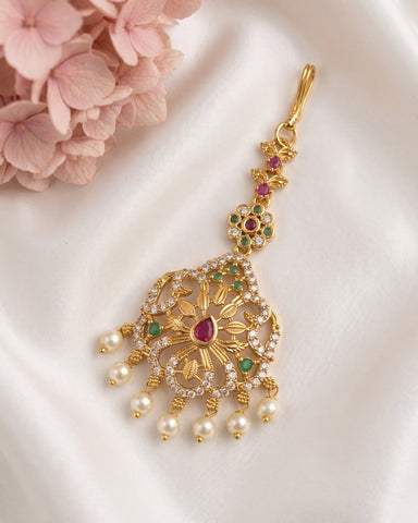 An ornate gold-toned Indian bridal maang tikka with a central ruby cabochon, green emerald accents, and dangling white faux pearls, displayed on a white satin fabric with pink flowers in the corner.