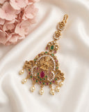 An ornate gold-toned traditional pendant or choker with intricate filigree, small clear stones, and red and green gemstone accents, displayed on white satin fabric with pink flowers in the corner.