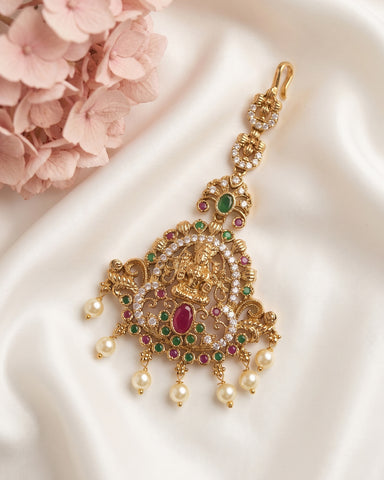 An ornate gold-toned traditional pendant or choker with intricate filigree, small clear stones, and red and green gemstone accents, displayed on white satin fabric with pink flowers in the corner.