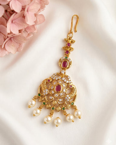 An ornate gold-toned traditional Indian jhumka earring decorated with red gemstones, clear stones, and dangling white faux pearls, displayed on a white fabric background with pink flowers in the corner.