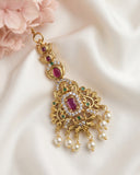 An ornate gold-toned traditional Indian pendant necklace with a large rectangular ruby-red central stone, smaller red stones, green accent stones, and dangling white pearl beads, displayed on a white satin fabric with pink flowers.