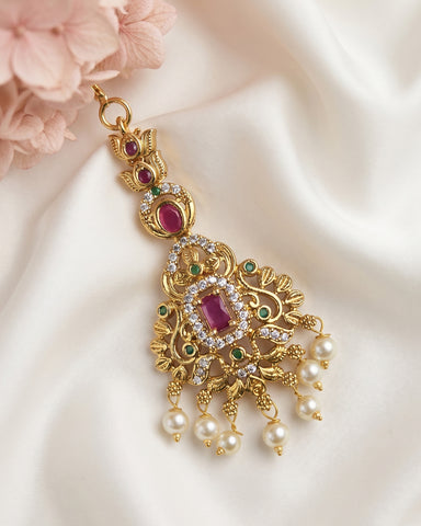 An ornate gold-toned traditional Indian pendant necklace with a large rectangular ruby-red central stone, smaller red stones, green accent stones, and dangling white pearl beads, displayed on a white satin fabric with pink flowers.