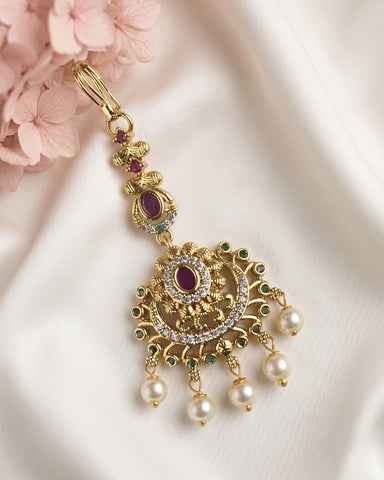 An ornate gold-toned traditional Indian choker necklace with a large pendant, featuring a central oval ruby stone, small green stones, clear crystals, and dangling white pearls, styled on white satin fabric with pink flowers.
