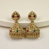 A pair of ornate gold-toned Indian jhumka earrings with red and green stone accents, displayed on a white curved riser against a light beige background.