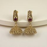 A pair of gold-plated Indian jhumka earrings with small red gemstone accents, displayed on a white curved stand against a light beige background.