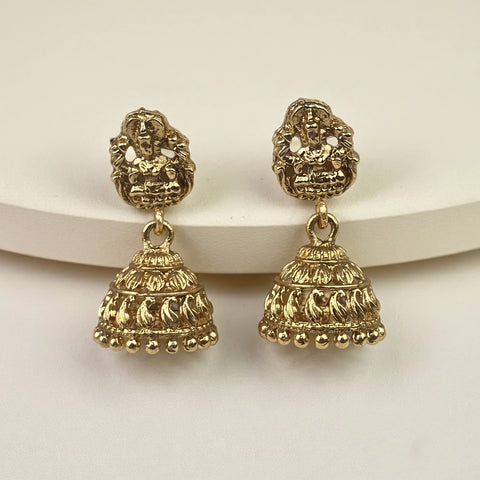 A pair of gold-toned Indian jhumka earrings with detailed filigree and beadwork, displayed on a white curved riser against a light beige background.