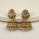 A pair of ornate gold-toned Indian jhumka earrings with detailed filigree work, small red and green gemstone accents, and dangling bell-shaped charms, displayed on a light cream background.