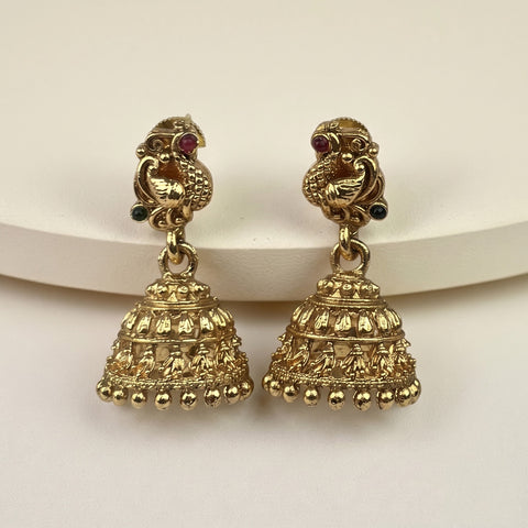 A pair of gold-plated Indian jhumka earrings with detailed filigree bell-shaped drops and peacock motifs on the posts, displayed on a white curved riser against a light beige background.