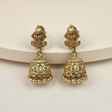 A pair of gold-toned Indian jhumka earrings with detailed filigree and a small deity figure at the top, displayed on a curved cream-colored surface against a light beige background.