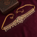 Gold-toned choker necklace with intricate peacock motif, green and red stone accents, and dangling pearl-like beads, displayed on a deep burgundy fabric with a carved wooden box.