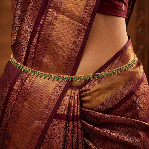 A close-up of a model wearing a gold-plated traditional Indian waist chain (bangle) embellished with small green stones, paired with a richly patterned maroon and gold silk saree.