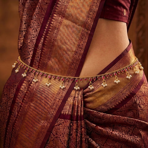 A close-up of a woman wearing a richly patterned bronze and burgundy saree with a gold-toned waist chain decorated with small red stones and white pearl-like drops.