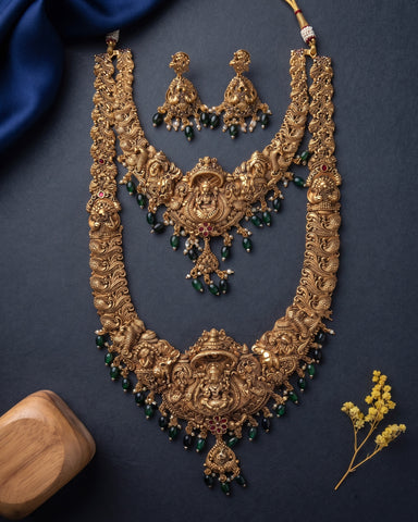 An ornate gold-toned traditional necklace set with matching earrings, decorated with green bead drops and small pink stone accents, arranged on a dark navy background with decorative fabric and dried flowers.