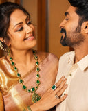 A close-up lifestyle shot of a couple wearing traditional Indian jewelry. The woman on the left, wearing a gold and pink saree, is adorned with a large gold and emerald green necklace, matching earrings, and a green square ring, with her hand resting on the man's chest.