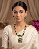 A woman wearing a gold and green jewelry set with a matching necklace and earrings, paired with a cream-colored sari. The jewelry features emerald-cut green stones set in gold.