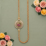 A gold-toned traditional Indian necklace with a decorative circular pendant featuring a pink peacock motif and white stones, displayed on a sage green background with decorative pink and yellow roses.