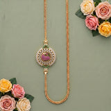 A gold-toned traditional Indian necklace with a central paisley motif, decorated with white stones and green and pink accents, displayed on a sage green background with decorative roses.