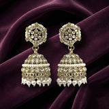 A pair of ornate gold-toned jhumka earrings decorated with white pearls and clear rhinestones, displayed on a draped deep purple velvet fabric.