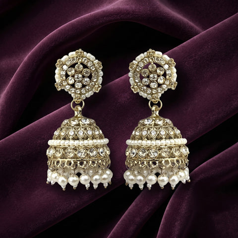 A pair of ornate gold-toned jhumka earrings decorated with white pearls and clear rhinestones, displayed on a draped deep purple velvet fabric.