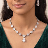 A close-up of a woman wearing a silver-toned necklace with diamond-like floral clusters and a teardrop pendant, along with matching drop earrings and a dark green velvet top.