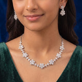 A close-up of a woman wearing a silver-toned floral necklace and matching flower-shaped drop earrings against a dark blue background.