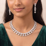 A close-up of a model wearing a silver-toned diamond necklace and matching drop earrings, paired with a dark green velvet top.