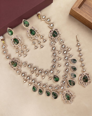 A traditional Indian jewelry set featuring a necklace, earrings, and a dangling charm in silver-tone metal with green teardrop stones and clear rhinestones, displayed on a beige surface.