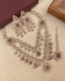 An ornate Indian ethnic jewelry set featuring a necklace, earrings, and a dangling maang tikka in silver-tone metal with teardrop-shaped brown gemstones and clear stones, displayed on a beige surface.