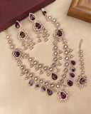 A traditional Indian jewelry set featuring a silver-tone necklace, earrings, and a maang tikka with deep purple teardrop stones and clear rhinestones, displayed on a beige surface.