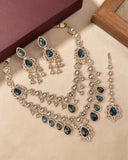 A traditional Indian jewelry set featuring a silver-tone necklace, earrings, and a dangle, embellished with silver stones and deep blue teardrop gemstones, displayed on a beige surface.