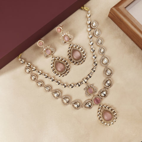 A gold-plated Indian ethnic jewelry set featuring a necklace, earrings, and a bracelet, decorated with pink stone accents and clear crystals, displayed on a beige surface.