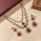 A traditional Indian jewelry set featuring a silver-toned necklace, earrings, and a matching bracelet, embellished with clear stones and deep red teardrop gemstones, displayed on a beige surface.