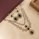 Gold-toned Indian ethnic jewelry set featuring a necklace, earrings, and bracelet with green teardrop and oval stones, arranged on a beige surface with decorative blocks in the background.