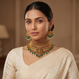 A woman wearing an ornate gold-toned traditional necklace with green and white stone accents, matching dangling earrings, and a cream-colored blouse against a warm beige indoor background.