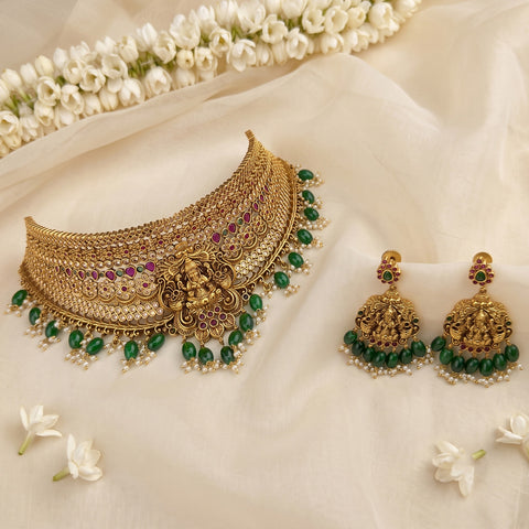 Gold-toned traditional Indian jewelry set featuring a choker necklace and matching dangle earrings, decorated with green beads, white pearls, and small purple stones, arranged on cream fabric with white flowers.