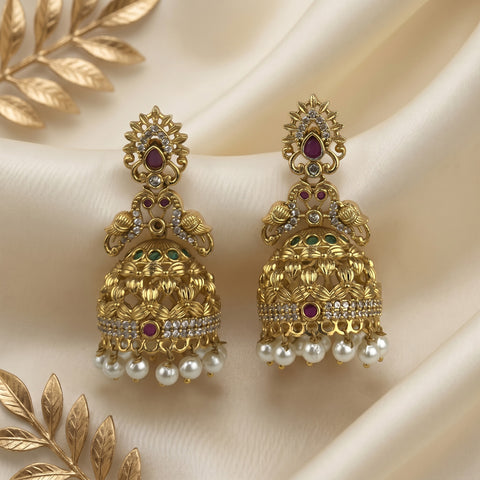 A pair of ornate gold-toned jhumka earrings with white pearls, small green and red stones, and clear crystal accents, displayed on draped cream satin fabric with decorative gold leaf elements.