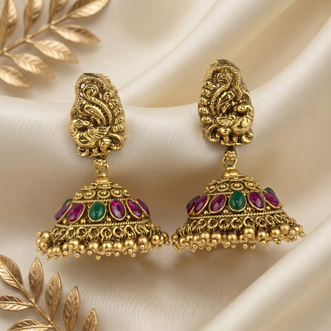 A pair of ornate gold-toned jhumka earrings with detailed filigree, pink and green stone accents, and dangling bead borders, displayed on draped cream satin fabric with decorative gold leaf elements.
