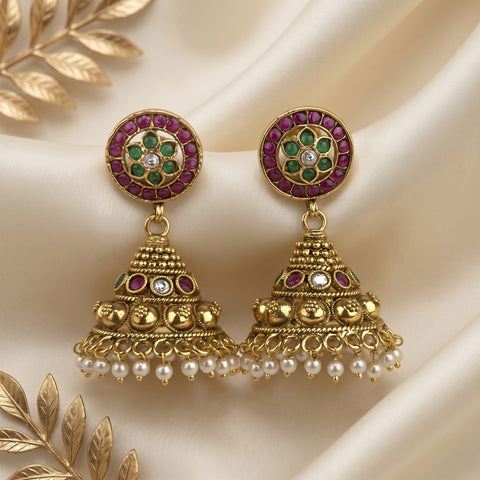 A pair of ornate gold-toned Indian jhumka earrings with circular studs featuring green and purple stones, dangling bell-shaped charms with gold filigree, small red stone accents, and rows of white faux pearls, displayed on draped cream satin fabric with decorative gold leaf elements.