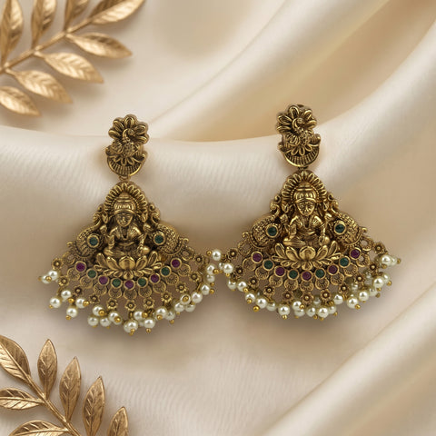 A pair of ornate gold-toned traditional Indian jhumka earrings with detailed Ganesha motifs, small multicolor gemstones, and rows of white faux pearls, displayed on draped cream satin fabric with decorative gold leaf props.