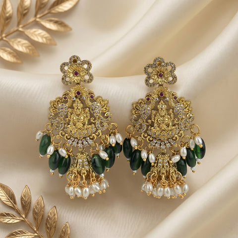 A pair of ornate gold-toned chandelier earrings with green stone drops, white pearl-like beads, and small red gemstone accents, displayed on draped cream fabric with decorative gold leaf elements.