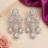 A pair of silver-toned drop earrings with clear teardrop and round gemstones, arranged on a beige surface with a pink rose, cream box corner, and magenta satin fabric.