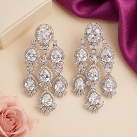 A pair of silver-toned drop earrings with clear teardrop and round gemstones, arranged on a beige surface with a pink rose, cream box corner, and magenta satin fabric.