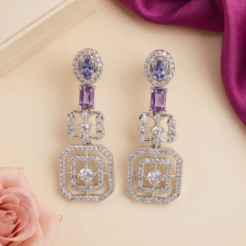 A pair of silver-tone drop earrings with oval blue gemstones, rectangular purple stones, and square crystal pavé settings, displayed on a beige surface with a pink rose and purple fabric accents.