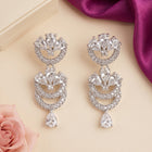 A pair of silver-tone drop earrings adorned with clear crystals, arranged on a light beige background with decorative pink rose petals and a purple satin ribbon.