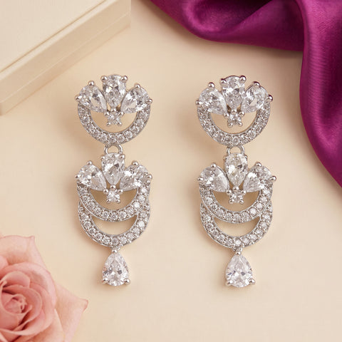 A pair of silver-tone drop earrings adorned with clear crystals, arranged on a light beige background with decorative pink rose petals and a purple satin ribbon.