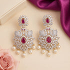 A pair of ornate gold and silver-toned dangle earrings decorated with clear pavé stones, red teardrop rubies, and white pearl-like drops, styled on a beige surface with a pink rose and purple satin fabric.